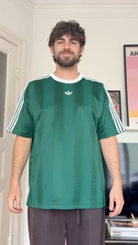 Man with dark hair and beard wearing a green Adidas sports shirt with white stripes, standing indoors in a room with framed art.