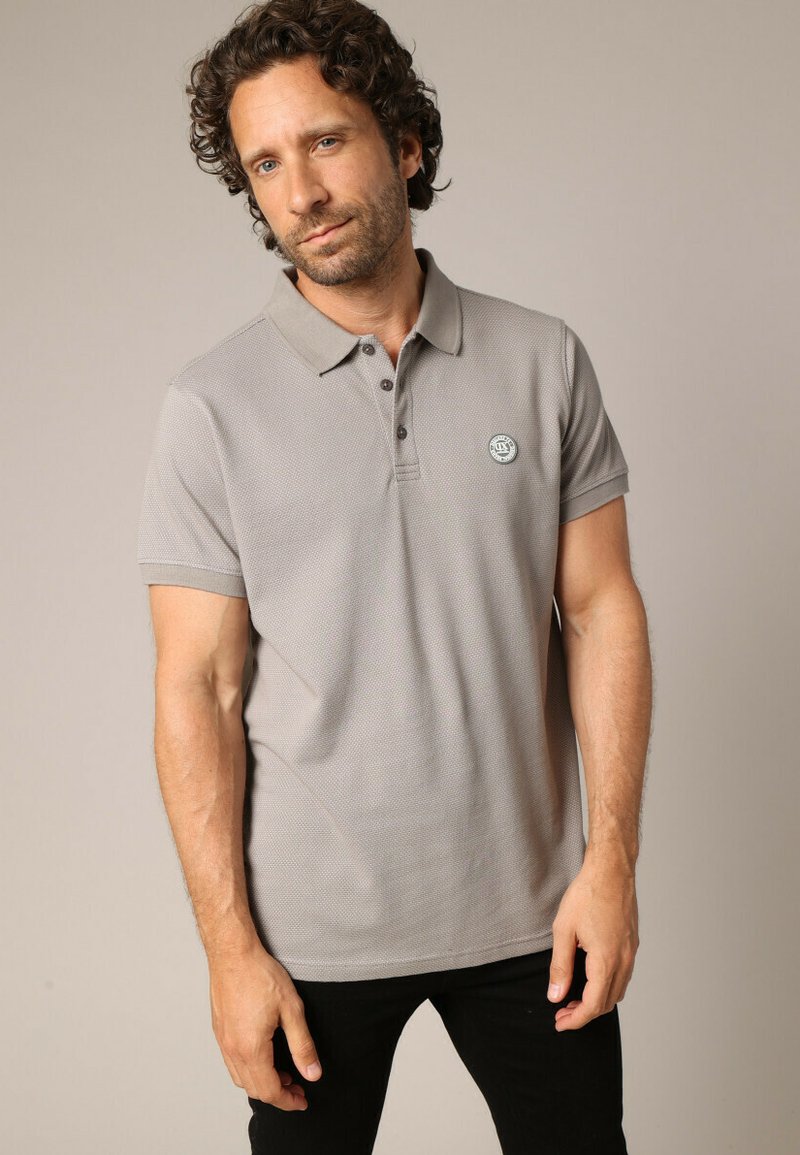 Grijze polo met een gestructureerde stof, een knoopsluiting met twee knopen en een rond logo-opdruk op de linkerkant van de borst, gecombineerd met zwarte broek.