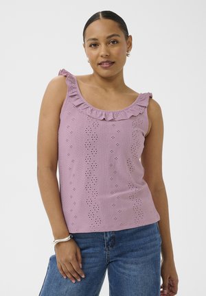 Femme portant un haut sans manches mauve en tissu ajouré avec un col à volants et un jean bleu, debout devant un fond blanc uni.