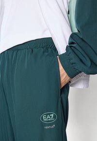 Σκούρο μπλε αθλητικό παντελόνι με ελαστική μέση, που διαθέτει λογότυπο και μαλακό, ελαφρύ ύφασμα. Το συνοδευτικό μπουφάν είναι λευκό.
