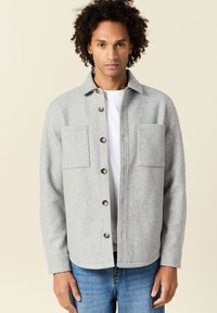 Veste boutonnée en laine grise avec deux poches poitrines, coupe droite et ourlet arrondi. Les boutons sont marron foncé. Portée par-dessus un t-shirt blanc.