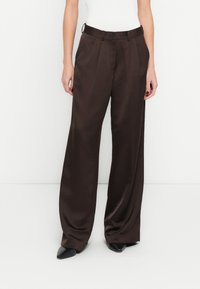 Pantalon marron à jambes larges en tissu lisse, avec un devant plissé et une ceinture à passants. Porté avec des chaussures noires pointues.