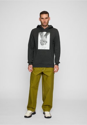 Homme portant un sweat à capuche gris foncé avec un motif de mains se tenant et le texte « Trust », un pantalon large vert olive et des baskets blanches, debout devant un fond gris.