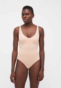 Nude badedrakt med dyp V-hals, tykke stropper og sømløst design. Stoffet har en glatt, matt overflate med en tettsittende silhuett.