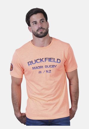 Homme barbu portant un T-shirt à manches courtes pêche avec le texte « RUCKFIELD MAORI RUGBY N.Z », debout devant un fond clair uni.