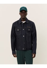 Giacca di jeans scuro con taglio classico, bottoni argentati e due tasche sul petto; abbinata a un cappellino teal con scritte ricamate.