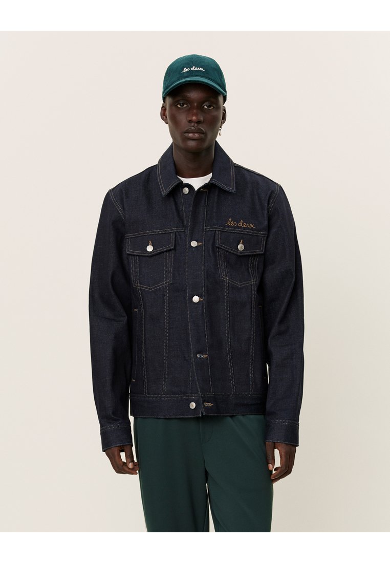 Giacca di jeans scuro con taglio classico, bottoni argentati e due tasche sul petto; abbinata a un cappellino teal con scritte ricamate.