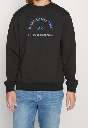 Person trägt schwarzen Pullover mit mehrfarbigem Text "KARL LAGERFELD PARIS 21 RUE ST-GUILLAUME", kombiniert mit hellblauen Jeans.