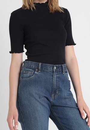 Vrouw die een zwart geribbeld kortemouwen top draagt met geschulpte randen en een hooggetailleerde blauwe spijkerbroek.