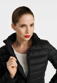 Czarna ocieplana kurtka z wysokim kołnierzem, z poziomymi pikowanymi liniami i zapięciem na zamek. Model trzyma otwarty kołnierz. Biała koszula pod spodem.