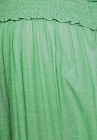 Tecido verde com uma textura suave e ligeiramente brilhante. Apresenta uma cintura elástica pregueada com borda em forma de ondas. Aparência leve e arejada.