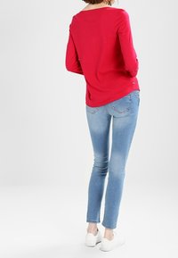 Top rojo de manga larga, con cuello redondo, combinado con jeans ajustados de un azul claro. Zapatillas blancas con diseño perforado completan el conjunto.