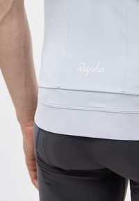 Zbliżenie osoby noszącej jasnoniebieską koszulkę kolarską i czarne wyściełane spodenki kolarskie, pokazujące logo Rapha na dolnej części pleców koszulki.