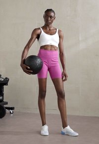 Un outfit sportiv format dintr-un top alb scurt și pantaloni scurți roz cu talie înaltă, completat de adidași albi și blue. Ținând o minge medicinală neagră.