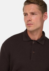 Brun, texturerad pikétröja med kort krage, tre bruna knappar och en figurnära design, presenterad på en manlig modell mot en neutral bakgrund.