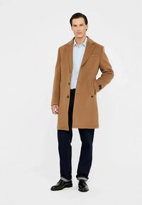 Brauner Wollmantel mit abgesteppten Revers, Ein-Knopf-Verschluss, seitlichen Taschen und maßgeschneiderter Passform, kombiniert mit einem hellblauen Hemd und dunkler Jeans.