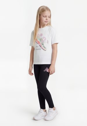 Ragazza giovane che indossa una t-shirt bianca New Balance con una grafica di sneaker e fiori, leggings neri con logo rosa e sneaker bianche.