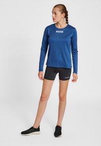 Marinblå långärmad atletisk topp med rund hals och logotyp, kombinerad med åtsittande svarta shorts och svarta sneakers. Slät textur, fuktavledande tyg.