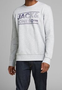 Felpa grigia in cotone con polsini e orlo a costine. Presenta il logo "JACK & JONES" in viola sul petto. Abbinata a jeans scuri.
