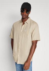 Chemise beige à manches courtes avec un bouton et une poche, en matière semblable au lin. Portée avec un jean bleu, une casquette noire et des lunettes de soleil foncées.
