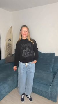 Μαύρο φούτερ με κουκούλα και γραφικό σχέδιο, ανοιχτό μπλε φαρδιά τζιν και σκούρα αθλητικά παπούτσια. Στο φόντο διακρίνεται ένας πράσινος καναπές και μια σανίδα του σερφ.