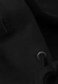 Svart stoff med en jevn tekstur, med en sølvfarget metallring og en snor, liten brodert detalj synlig.