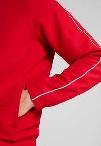 Rote Sportjacke mit glatter Textur, langen Ärmeln, Taschen an den Seiten und weißen Paspeln entlang der Ärmel.