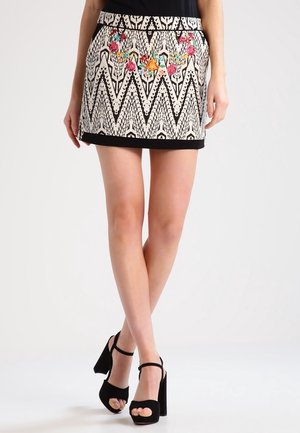 Kobieta w mini spódniczce w czarno-biały wzór zygzaków ozdobionej kolorową kwiatową haftem oraz czarnych sandałach na wysokim obcasie z odkrytymi palcami.