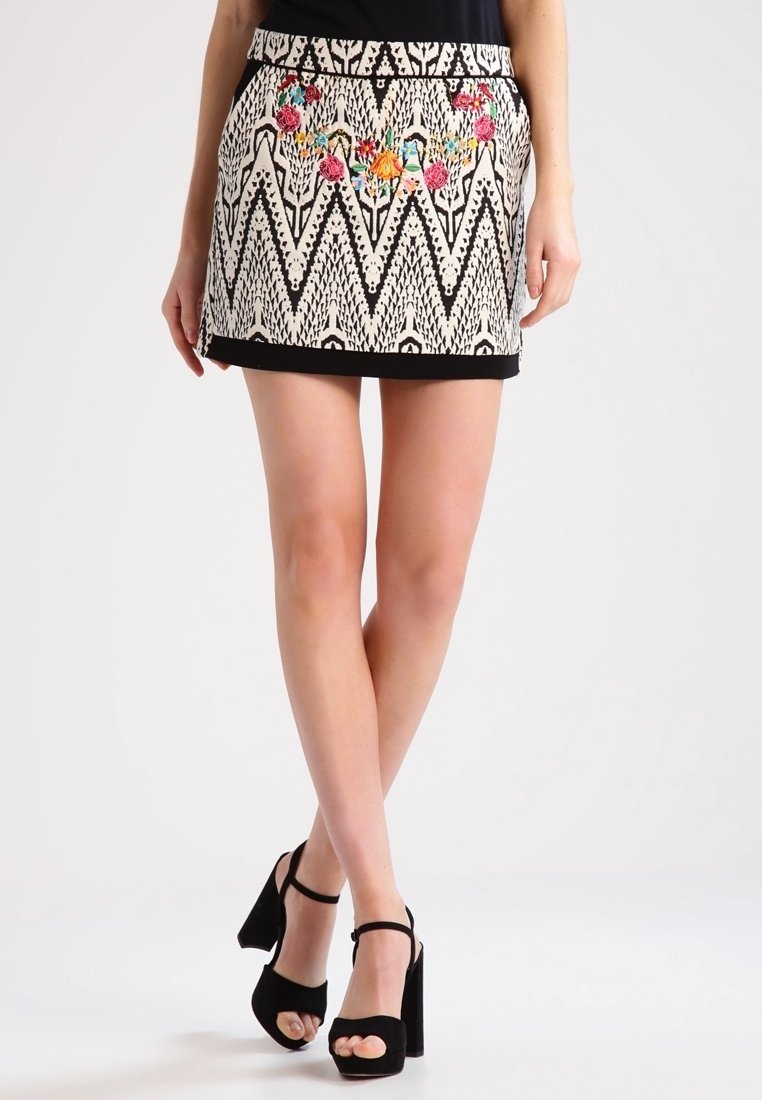 Femme portant une mini-jupe à motif zigzag noir et blanc avec une broderie florale colorée et des sandales noires à talons hauts et bout ouvert.