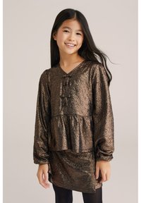 Brūna, strāvojoša, garām piedurknēm kleita ar pepluma jostu un trim dekoratīvām pogām pie kakla. Stilizēta ar melnajām zeķubiksēm.