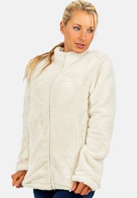 Femme portant une veste crème en tissu pelucheux avec fermeture éclair, regardant sur le côté, les mains au bas de la veste sur fond blanc.