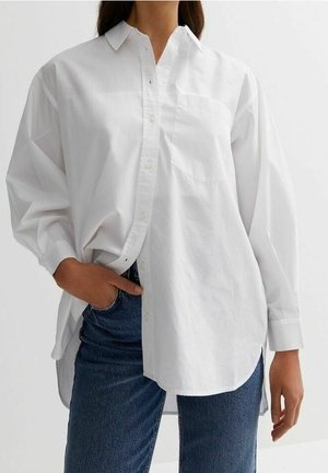 Vrouw draagt een oversized wit overhemd met lange mouwen en een blauwe spijkerbroek, hand in de zak, staande tegen een effen achtergrond.