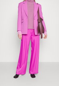 Blazer rose à revers crantés, associé à un col roulé côtelé rose clair et un pantalon large en satin rose vif. Sac à épaule marron.