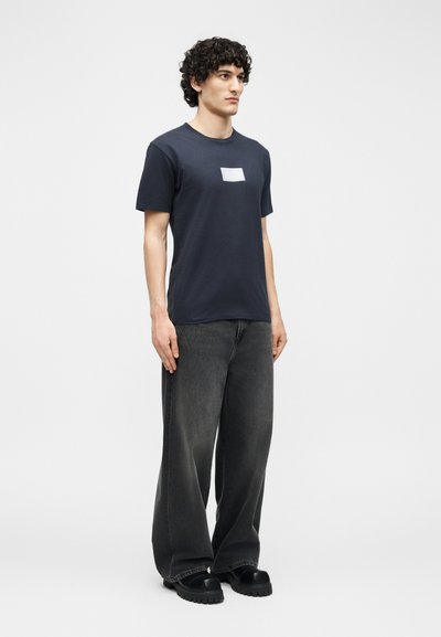 Άνδρας με σγουρά μαλλιά, φορώντας μπλε ναυτικό T-shirt με λευκή επιγραφή, φαρδιά μαύρα τζιν και μαύρα χοντρά παπούτσια, στέκεται μπροστά σε απλό φόντο.