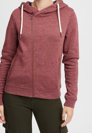 Personne portant un sweat à capuche bordeaux zippé avec des cordons blancs et un pantalon vert foncé, debout devant un fond clair uni.