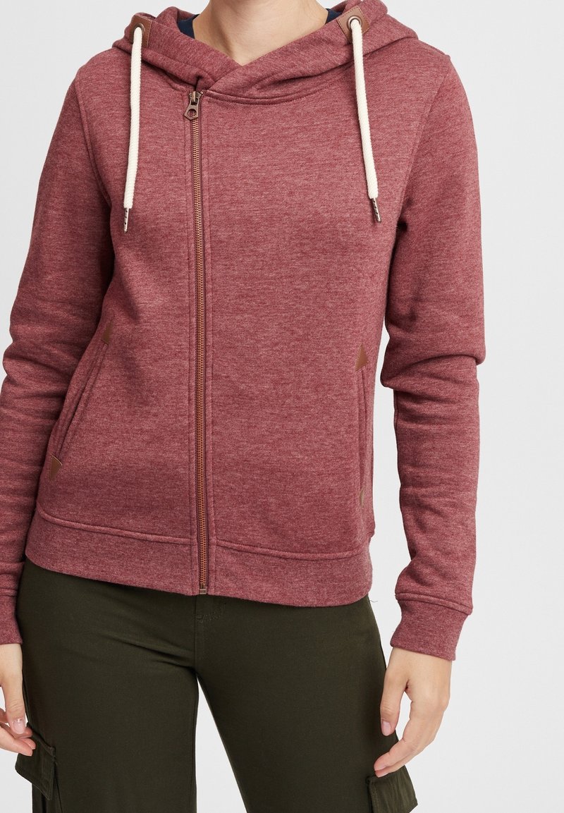 Personne portant un sweat à capuche bordeaux zippé avec des cordons blancs et un pantalon vert foncé, debout devant un fond clair uni.