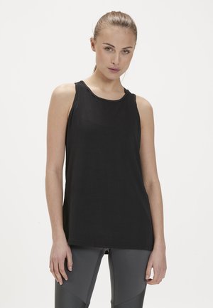 Kvinde med lys hud og håret bundet tilbage, iført en sort ærmeløs top og grå leggings mod en ensartet hvid baggrund.