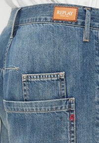 Ανοιχτό μπλε τζιν παντελόνι με τετράγωνη πίσω τσέπη, που διαθέτει πορτοκαλί δερμάτινη ετικέτα με λογότυπο και κόκκινη ραφή λεπτομέρεια στην άκρη της τσέπης.