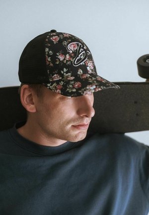 Crna cvjetna kapa s logotipom, koju nosi osoba uz svijetli zid. Drži skateboard, obučena u tamnu duksericu.