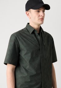 Korte mouwen shirt in donkergroen, met een geometrisch patroon, knoopsluiting, borstzak met rits en subtiele branding.