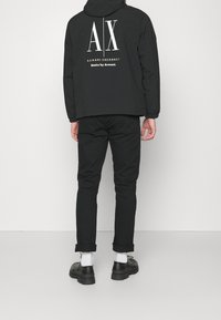 Svart huvan jacka med en vit grafisk "AX" logotyp och text på baksidan. Matchad med svarta byxor och svarta skor med vita strumpor.