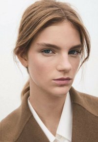 Zbliżenie osoby o jasnobrązowych włosach i niebieskich oczach, ubranej w brązowy płaszcz i białą koszulę. Skóra wygląda na gładką, z naturalnym makijażem.