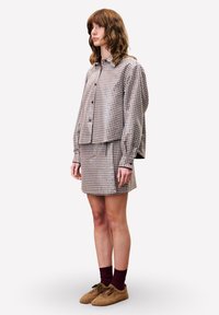 Vrouw staand in een bruin geruit oversized overhemd, bijpassend minirok, bordeauxrode sokken en beige pluizige schoenen, kijkend naar links.