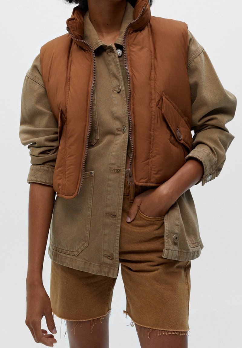 Brown puffer vest over a khaki shirt jacket, featuring a zip closure and front pockets. Matching brown shorts with frayed edges.