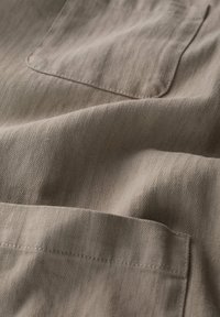 Tessuto beige con un sottile motivo a spina di pesce, caratterizzato da una tasca visibile e da una consistenza liscia, catturato in dolci pieghe e ombre leggere.