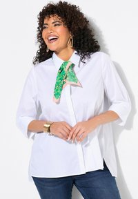 Camicia bianca con maniche a tre quarti, caratterizzata da una sciarpa a fantasia verde e rosa legata al colletto. Indossata con jeans scuri.