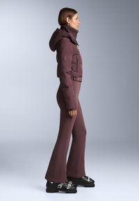 Vrouw in een bordeauxrode gewatteerde jas met capuchon, bijpassende uitlopende broek en zwarte skischoenen, staand in profiel tegen een effen grijze achtergrond.