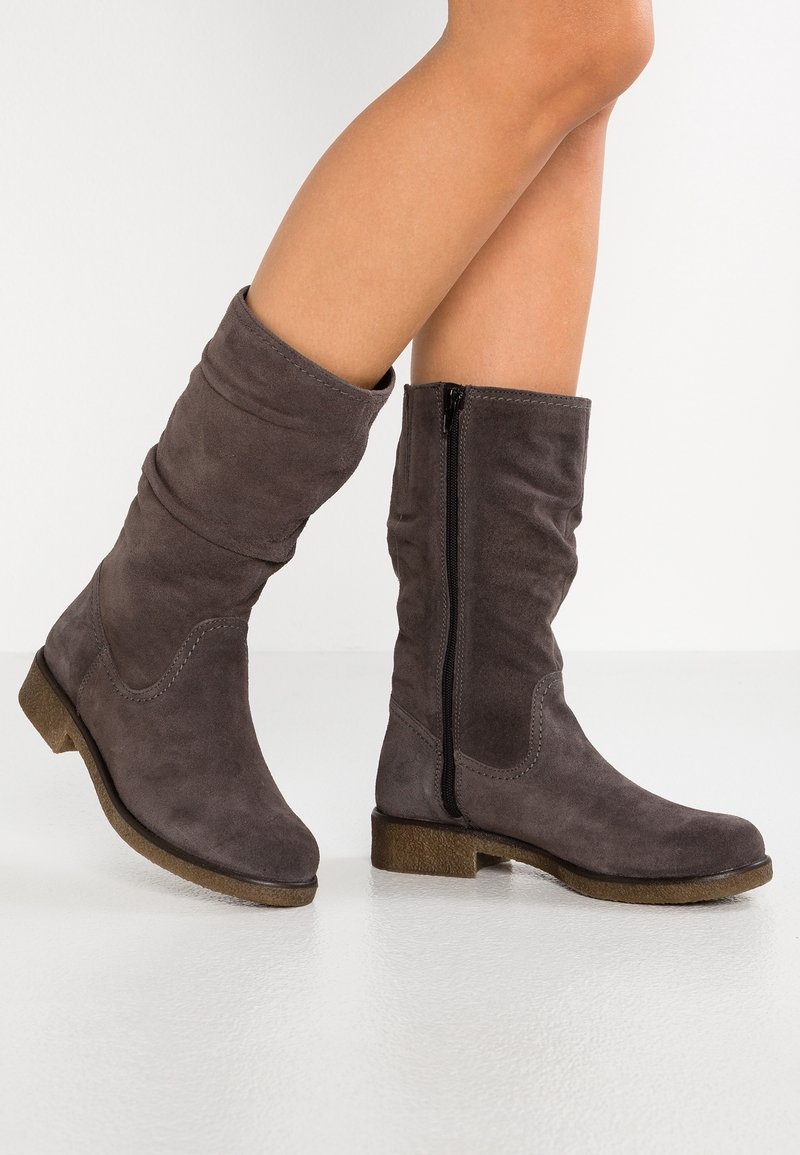 Femme portant des bottes en daim gris mi-mollet avec fermetures éclair latérales et talons bas en caoutchouc marron sur un sol blanc.
