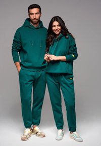 Sudaderas verdes y pantalones a juego hechos de una tela suave. Cada prenda cuenta con una capucha ajustable y puños elásticos. El calzado presenta tonos claros y neutros.