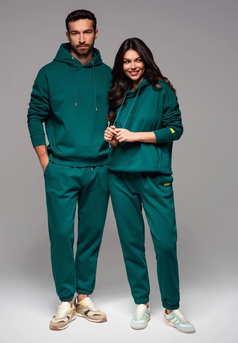 Sudaderas verdes y pantalones a juego hechos de una tela suave. Cada prenda cuenta con una capucha ajustable y puños elásticos. El calzado presenta tonos claros y neutros.